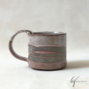 May include: A ceramic mug with a cylindrical body and a curved handle. The exterior features a speckled glaze with horizontal bands of muted pink, grey, and brown tones. The surface has a textured appearance with visible colour transitions.