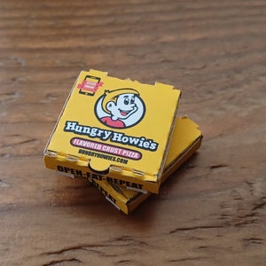 May include: Two yellow pizza boxes stacked on top of each other. The top box has the words "Hungry Howie's" and "Flavored Crust Pizza" printed on it. The bottom box has the words "Open-Eat-Repeat" printed on it.