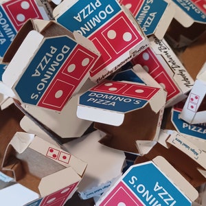 Pu&ograve; includere: Un mucchio di scatole per pizza Domino's di vari colori, tra cui blu, rosso e bianco. Le scatole sono impilate l'una sull'altra e alcune sono aperte. Le scatole hanno il logo Domino's e le parole "Domino's Pizza" stampate su di esse. Le scatole sono fatte di cartone e hanno un interno bianco.