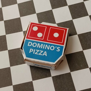Pu&ograve; includere: Una scatola per pizza Domino's bianca e blu con il logo Domino's e le parole "Domino's Pizza" stampate su di essa. La scatola &egrave; appoggiata su una superficie a scacchi bianchi e neri.