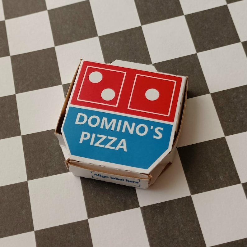 Pu&ograve; includere: Una scatola per pizza Domino's bianca e blu con il logo Domino's e le parole "Domino's Pizza" stampate su di essa. La scatola &egrave; appoggiata su una superficie a scacchi bianchi e neri.