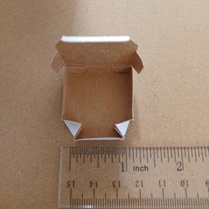 Pu&ograve; includere: Una piccola scatola di cartone marrone vuota con bordi bianchi. La scatola &egrave; aperta e l'interno &egrave; visibile. La scatola &egrave; larga circa 2,5 cm e lunga 2,5 cm.