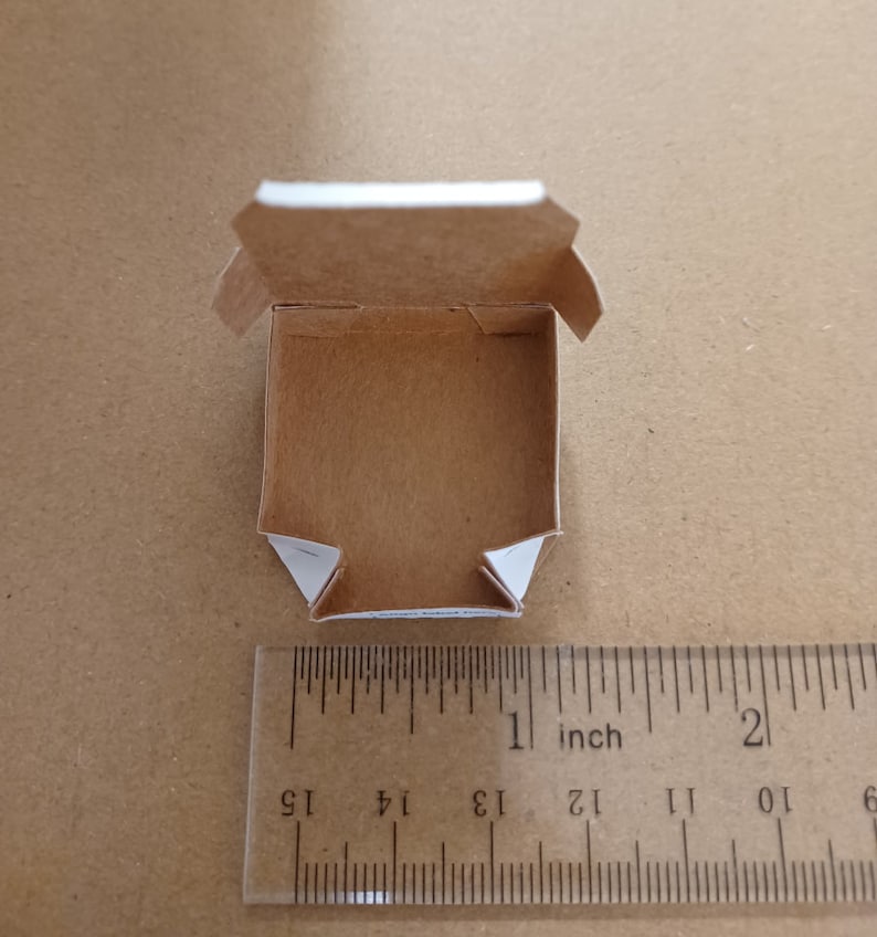 Pu&ograve; includere: Una piccola scatola di cartone marrone vuota con bordi bianchi. La scatola &egrave; aperta e l'interno &egrave; visibile. La scatola &egrave; larga circa 2,5 cm e lunga 2,5 cm.