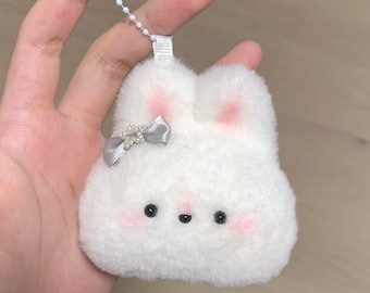 Könnte beinhalten: Ein weißer, flauschiger Hasen-Schlüsselanhänger mit einer silbernen Schleife und Perlen-Akzenten. Der Hase hat rosa Wangen und schwarze Augen.