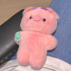 K&ouml;nnte beinhalten: Ein rosa Pl&uuml;schschweinchen mit einer lila Schleife auf dem Kopf. Das Schweinchen hat eine kleine, rosa Schnauze und schwarze Augen. Das Spielzeug sitzt auf dem Scho&szlig; einer Person.