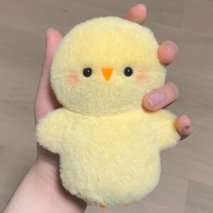 K&ouml;nnte beinhalten: Ein gelbes Pl&uuml;sch-K&uuml;kenspielzeug mit orangefarbenem Schnabel und schwarzen Augen. Das K&uuml;ken sitzt in einer Hand.