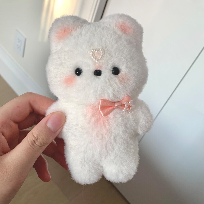 K&ouml;nnte beinhalten: Ein wei&szlig;er Pl&uuml;schb&auml;r mit rosa Wangen und einer rosa Schleife. Der B&auml;r hat schwarze Augen und ein Perlenherz auf der Stirn.