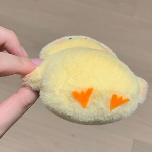 K&ouml;nnte beinhalten: Ein gelbes Pl&uuml;sch-K&uuml;kenspielzeug mit orangefarbenen Filzf&uuml;&szlig;en. Das K&uuml;ken liegt auf dem R&uuml;cken mit den F&uuml;&szlig;en in der Luft.