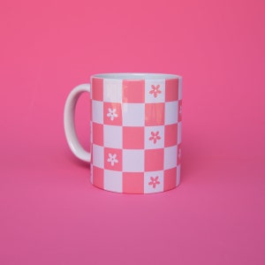 Puede incluir: Una taza de cerámica a cuadros rosa y blanco con un diseño floral. La taza tiene un asa blanca.