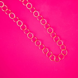 Puede incluir: Un collar de cadena de oro con eslabones grandes y redondos. La cadena está sobre un fondo rosa brillante.