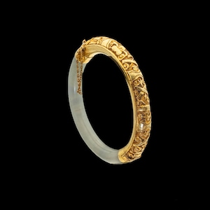 May include: A hinged bangle bracelet featuring a translucent, pale gray stone half-circle joined to a curved, ornate gold-tone metal section. The metal portion displays intricate, raised floral and scrollwork patterns and includes a thin safety chain.