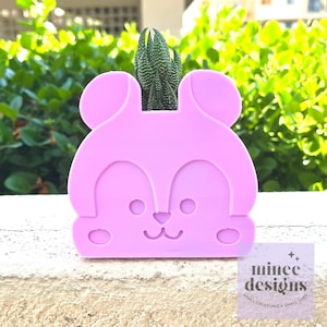 May include: A light purple, 3D printed planter shaped like a cartoon animal with large ears, a small nose, and two eyes. A small succulent plant is growing in the planter.