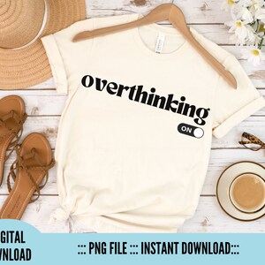 Könnte beinhalten: Ein weißes T-Shirt mit dem Text "overthinking ON" in Schwarz. Der Text ist in einer fetten, Retro-Schriftart. Das Shirt liegt flach auf einer weißen Holzoberfläche mit einem Paar Sandalen, einem Strohhut und einer Tasse Kaffee.