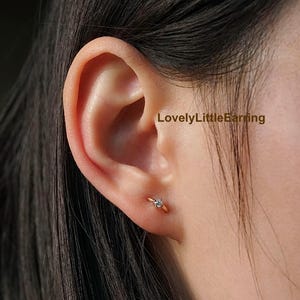 May include: A close-up of a person's ear with a small gold hoop earring. The earring features a tiny, sparkling gemstone. The text "LovelyLittleEarring" is visible in the image.