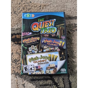 May include: A PC game case for "The Quest Trio" featuring three Mah Jong Quest games. The cover art includes colorful graphics, the game title, and the ESRB rating. The case is on a textured surface.