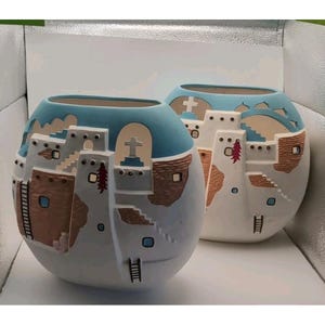 May include: Two spherical ceramic vases with a Southwestern design. The vases are white with blue rims and feature relief details of buildings, ladders, and crosses. The buildings have brown accents and red chili pepper details.