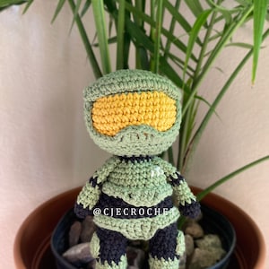 May include: A handmade crochet figure of a character from the Halo video game series. The figure is primarily green with yellow visor and black accents. The text "@CJECROCHE" is visible on the figure. The figure is in a brown pot with rocks and a green plant.