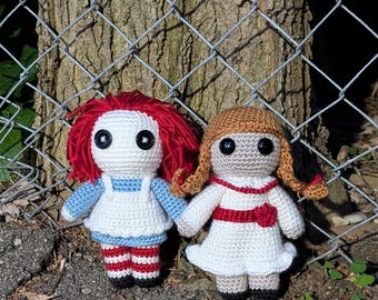 May include: Two handmade crochet dolls. One doll has red yarn hair, a white dress with blue accents, and red and white striped legs. The other doll has brown yarn hair, a white dress with a red belt, and black shoes. They are holding hands.