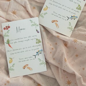 May include: Two watercolor menus with the word "Menu" at the top. Each menu features a border of green leaves and watercolor illustrations of food items like a carrot, fish, and lemon. The menus are on a pink fabric with gold star patterns.