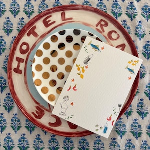 May include: A stack of ceramic plates with a decorative design and the text "HOTEL ROMA". A smaller plate with brown polka dots sits atop a light blue plate. A white card with illustrations of fish, bottles, and other designs is on top.