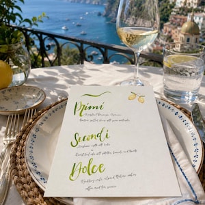 May include: A white rectangular menu card with green cursive headings Primi, Secondi, and Dolce, featuring a small lemon illustration. The menu is placed on a white plate with a blue floral rim, set on a woven placemat with a wine glass and water glass.