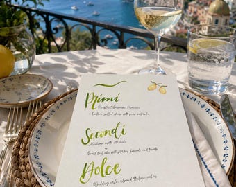 May include: A white rectangular menu card with green cursive headings Primi, Secondi, and Dolce, featuring a small lemon illustration. The menu is placed on a white plate with a blue floral rim, set on a woven placemat with a wine glass and water glass.