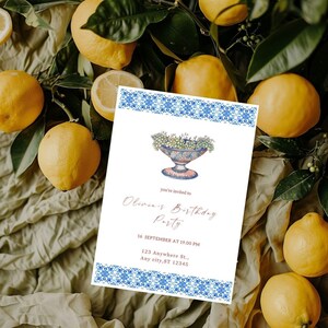 Könnte beinhalten: Eine weiße Einladungskarte mit einem blau-weißen Musterrahmen und einem floralen Design. Auf der Karte steht "Olivia's Birthday Party" mit Datum und Uhrzeit, umgeben von Zitronen und grünen Blättern.