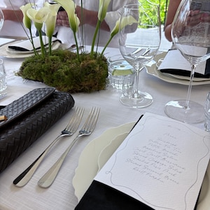 Puede incluir: Elegante mesa con mantel blanco, centro de mesa de calas y copas de vino. Incluye cubertería, un bolso de mano negro y una tarjeta de menú con texto manuscrito. La escena sugiere una experiencia gastronómica.