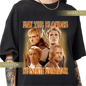 Può includere: Una t-shirt nera a maniche corte con una stampa grafica di quattro uomini biondi su uno sfondo di fulmini. Il testo "MAY THE BLONDES" appare in alto e "BE EVER IN YOUR FAVOR" in basso in lettere arancioni con un contorno scuro.