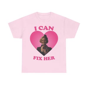 Può includere: Una t-shirt rosa chiaro con una grande grafica a cuore rosa scuro sul petto. All'interno del cuore c'è un ritratto fotografico di una persona con capelli biondi e una sciarpa a motivi. Il testo I CAN FIX HER è stampato in rosa scuro.