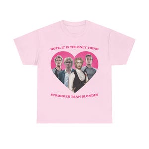 Puede incluir: Una camiseta de manga corta de color rosa claro con un gran gráfico de corazón rosa oscuro que contiene a cuatro hombres. Sobre el corazón está el texto "HOPE. IT IS THE ONLY THING" y debajo el texto "STRONGER THAN BLONDES" en letras mayúsculas.