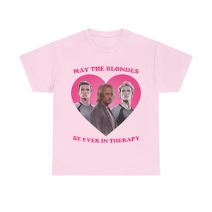 Può includere: Una t-shirt rosa chiaro con una grande grafica a cuore rosa scuro contenente una stampa fotografica di tre uomini. Sopra il cuore è presente il testo MAY THE BLONDES e sotto il cuore il testo BE EVER IN THERAPY in un carattere rosa grassetto.
