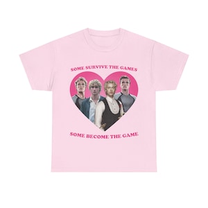 May include: A light pink t-shirt featuring a central dark pink heart graphic containing four male figures. The text "SOME SURVIVE THE GAMES" is printed above the heart, and "SOME BECOME THE GAME" is printed below the heart in dark pink lettering.