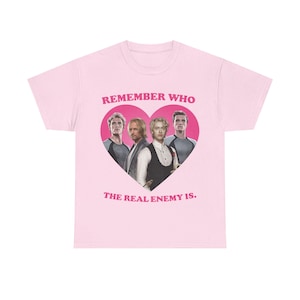 Può includere: Una t-shirt rosa chiaro con una grafica a cuore rosa scuro al centro che contiene quattro uomini. Sopra il cuore c'è il testo REMEMBER WHO e sotto THE REAL ENEMY IS. Gli uomini indossano vari abiti, tra cui gilet e camicie con texture.