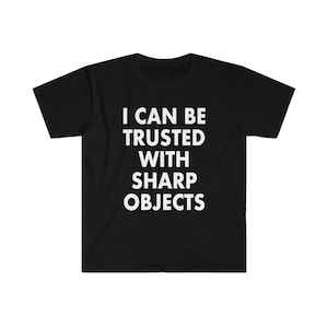 May include: Black t-shirt featuring the white text "I CAN BE TRUSTED WITH SHARP OBJECTS". The shirt has a crew neck and short sleeves. The text is centered on the front of the shirt.