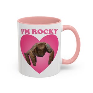 May include: White ceramic mug with a pink interior and handle. The mug features the text "I'M ROCKY" above a brown, textured sculpture inside a pink heart.