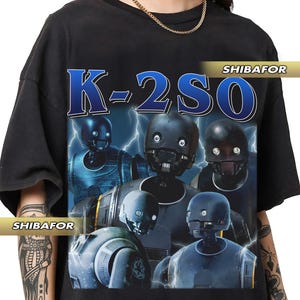 Puede incluir: Camiseta negra con un estampado gráfico de cinco robots del universo de Star Wars. Los robots están en varias poses y el texto "K-2SO" está impreso en azul en la camiseta.