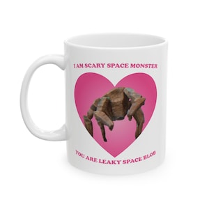 May include: White ceramic mug with a handle. The mug features a pink heart with a brown space monster illustration. Text on the mug reads "I AM SCARY SPACE MONSTER" and "YOU ARE LEAKY SPACE BLOB".