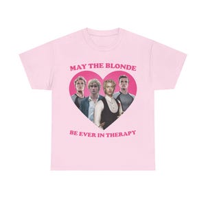 May include: A light pink short-sleeved t-shirt featuring a central pink heart graphic containing four blonde men. Above the heart is the text MAY THE BLONDE and below the heart is the text BE EVER IN THERAPY printed in a bold pink sans-serif font.
