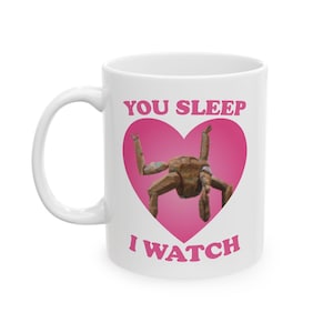 May include: White ceramic mug with the text "YOU SLEEP I WATCH" in pink. A wooden sculpture is inside a pink heart. The mug has a white handle and is suitable for hot or cold drinks.