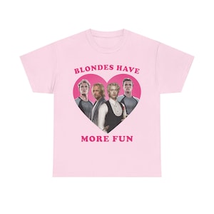 Può includere: T-shirt rosa chiaro con grafica a forma di cuore rosa con quattro uomini e la scritta "BLONDES HAVE MORE FUN". La t-shirt è a girocollo.