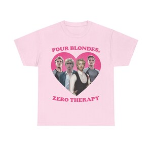 May include: A light pink short-sleeve t-shirt featuring a central graphic of a pink heart containing four blonde individuals. Above the heart is the text "FOUR BLONDES," and below the heart is the text "ZERO THERAPY" in a bold pink font.