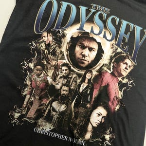 Pode incluir: Uma t-shirt preta com uma impressão gráfica contendo o texto THE ODYSSEY e CHRISTOPHER NOLAN. O design apresenta uma colagem de várias personagens e atores num estilo cinematográfico, com um retrato circular central rodeado por figuras.