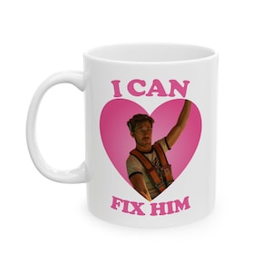 May include: White ceramic mug with a pink heart design. The words "I CAN" and "FIX HIM" are printed above and below a photo of a man wearing a harness and raising his arm.