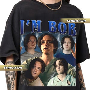Könnte beinhalten: Schwarzes T-Shirt mit einem Grafikdruck von vier Personen mit dem Text "I'M BOB" und "SHIBAFÖR" auf der Vorderseite.