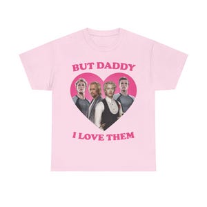 May include: A light pink short-sleeve t-shirt featuring a large dark pink heart graphic on the chest. Inside the heart are four men from The Hunger Games. Above the heart is the text BUT DADDY and below the heart is the text I LOVE THEM.