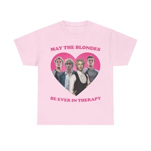 Può includere: Una t-shirt a maniche corte rosa chiaro con una grande grafica a cuore rosa scuro contenente un collage di quattro uomini biondi. Sopra il cuore è presente la scritta MAY THE BLONDES e sotto la scritta BE EVER IN THERAPY in grassetto rosa.