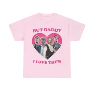 May include: A light pink short-sleeve t-shirt featuring a central graphic of a large dark pink heart containing four men. Above the heart is the text "BUT DADDY" and below the heart is the text "I LOVE THEM" in bold, dark pink capital letters.
