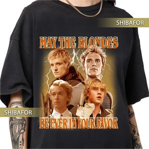 Puede incluir: Una camiseta negra de manga corta con un estampado gráfico de cuatro hombres rubios sobre un fondo de rayos. El texto "MAY THE BLONDES BE EVER IN YOUR FAVOR" está impreso en naranja con un borde negro sobre y bajo la imagen.