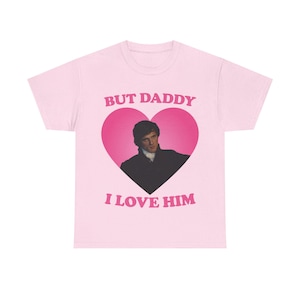 May include: A pale pink t-shirt with a pink heart design. Inside the heart is a man's image. The text "BUT DADDY" and "I LOVE HIM" are printed in pink above and below the heart.